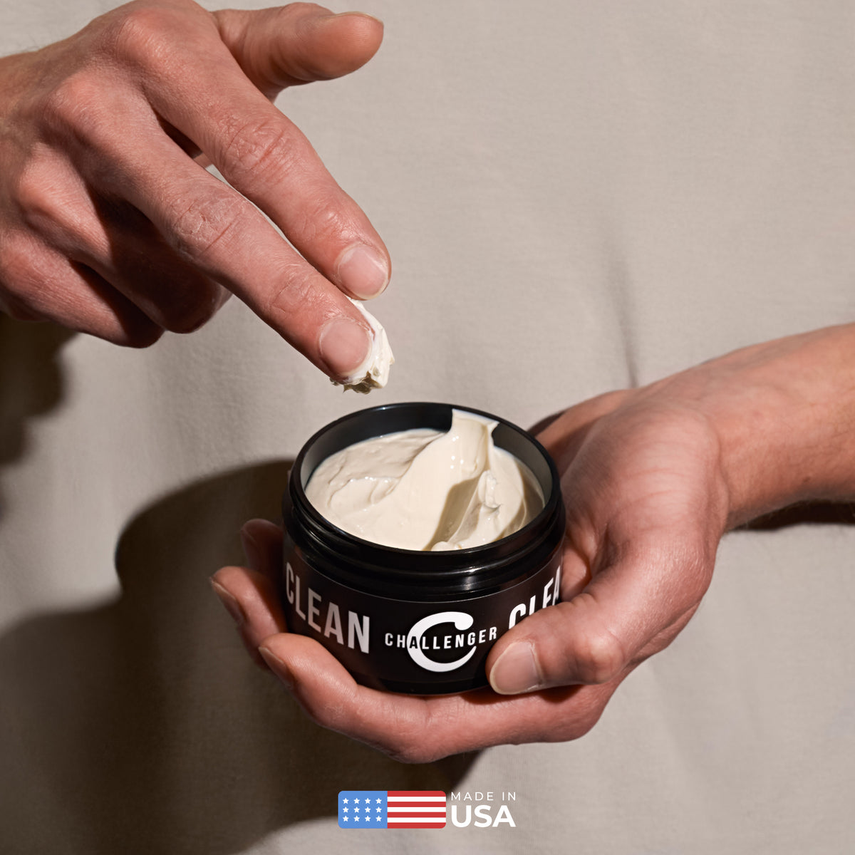 Hands holding an open jar of Challenger Clean pomade with a dab of white cream on the index finger.